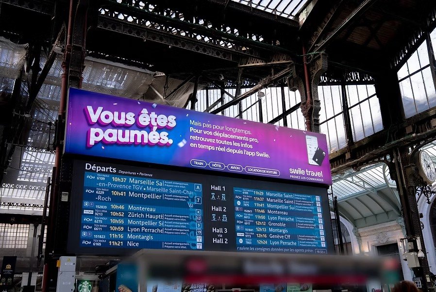 Affiche « Vous êtes paumés. » au-dessus du tableau des départs d'une gare.